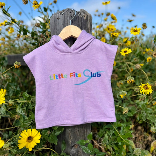 Purple hoodie with 'Little Fits Club' logo on a wooden post surrounded by yellow flowers.